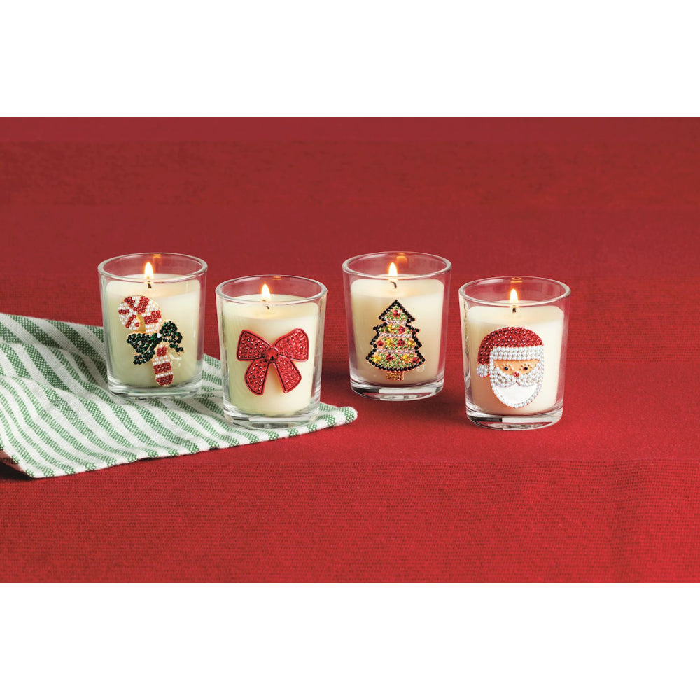 Four lit holiday-themed glass candle holders on a red textured background, with a green and white striped cloth under the first two candles. The designs include a candy cane, a red bow, a Christmas tree, and Santa Claus.