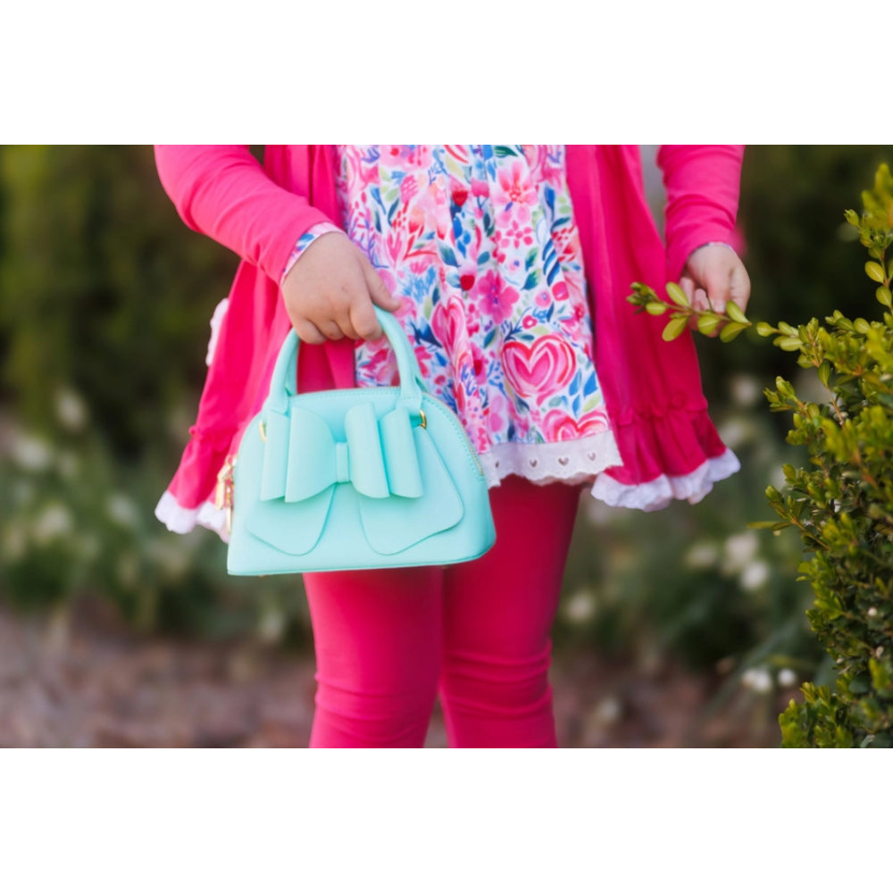 Child holding a light blue handbag with a pink jacket and floral dress in an outdoor setting