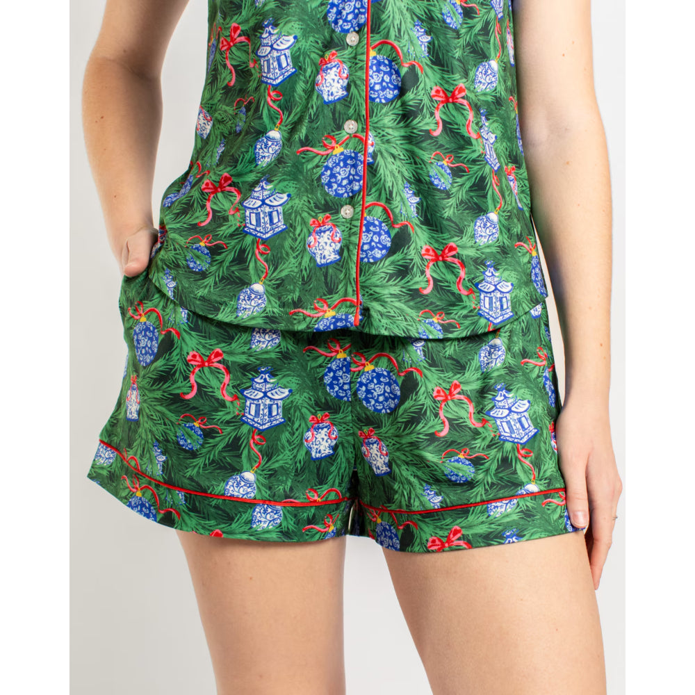 A person wearing a matching green pajama set with a Christmas print of pine branches, blue ornaments, and red bows, shown from the shoulders to the thighs against a light gray studio background.