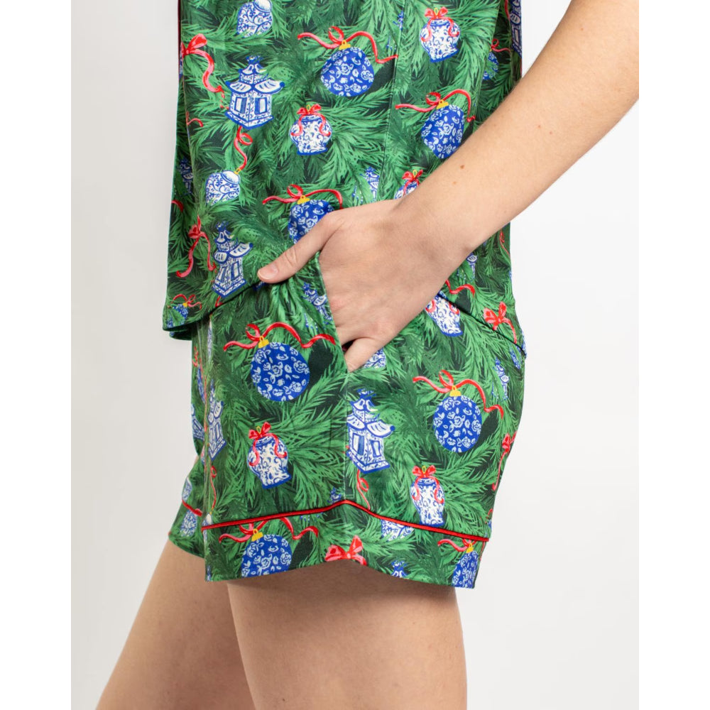 A close-up of a person wearing a green pajama set with a Christmas tree and ornament print, showing the shorts and part of the matching top, with one hand in the pocket.