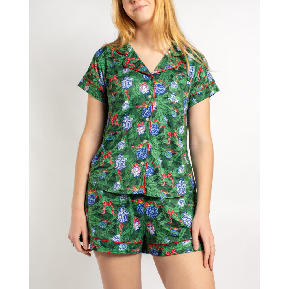 A woman wearing a two-piece pajama set with a green Christmas tree and gift print, featuring red piping on the collar, placket, cuffs, and shorts, against a plain light gray background.