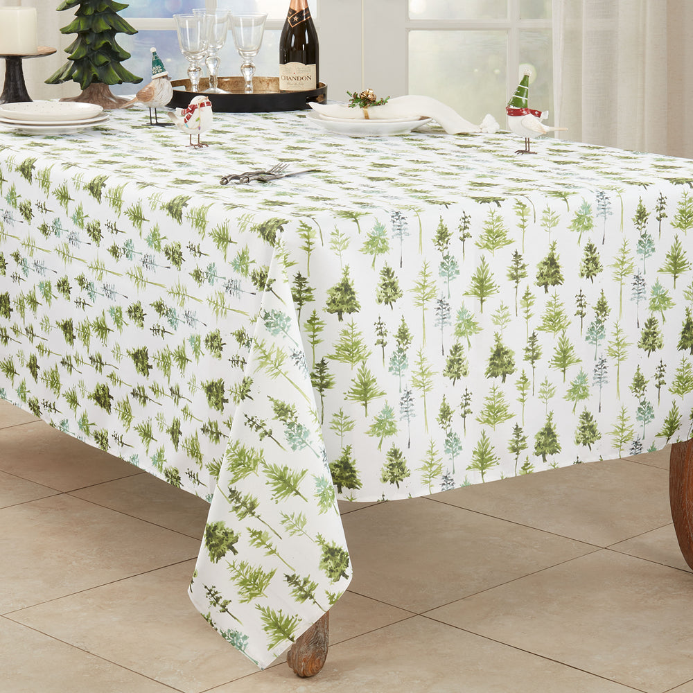A white tablecloth with a green and light blue pine tree pattern, shown draped over a dining table set with plates, glasses, and holiday decorations in a bright room.