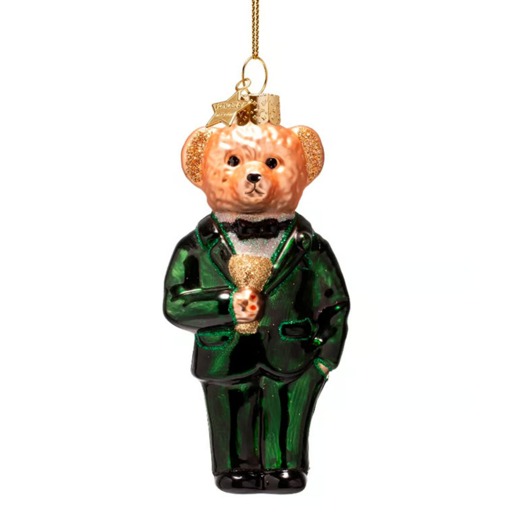 A glass Christmas ornament depicting a teddy bear dressed in a shiny emerald green tuxedo with a black bow tie and holding a gold trophy, suspended by a gold string against a white background.