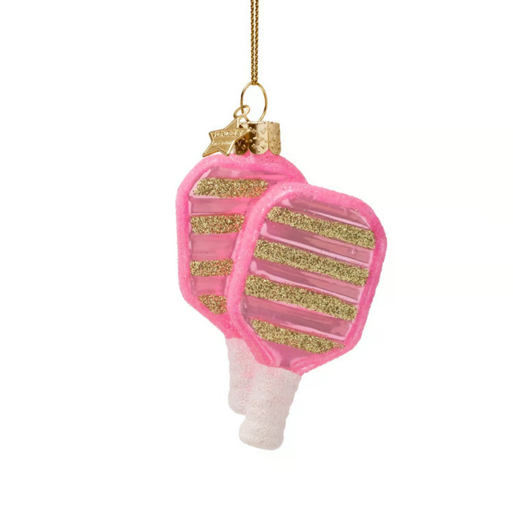 A pink and gold ornament shaped like a pair of pickleball rackets, hanging from a gold string against a plain white background.