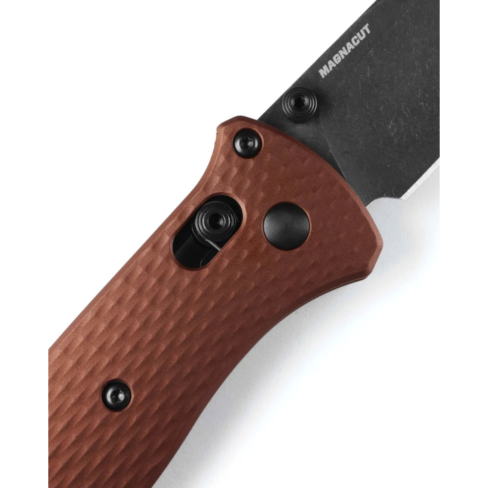 Close-up of a folding knife handle in textured reddish-brown metal with a black blade and black hardware, on a white background. The blade shows the MAGNA-CUT brand name.
