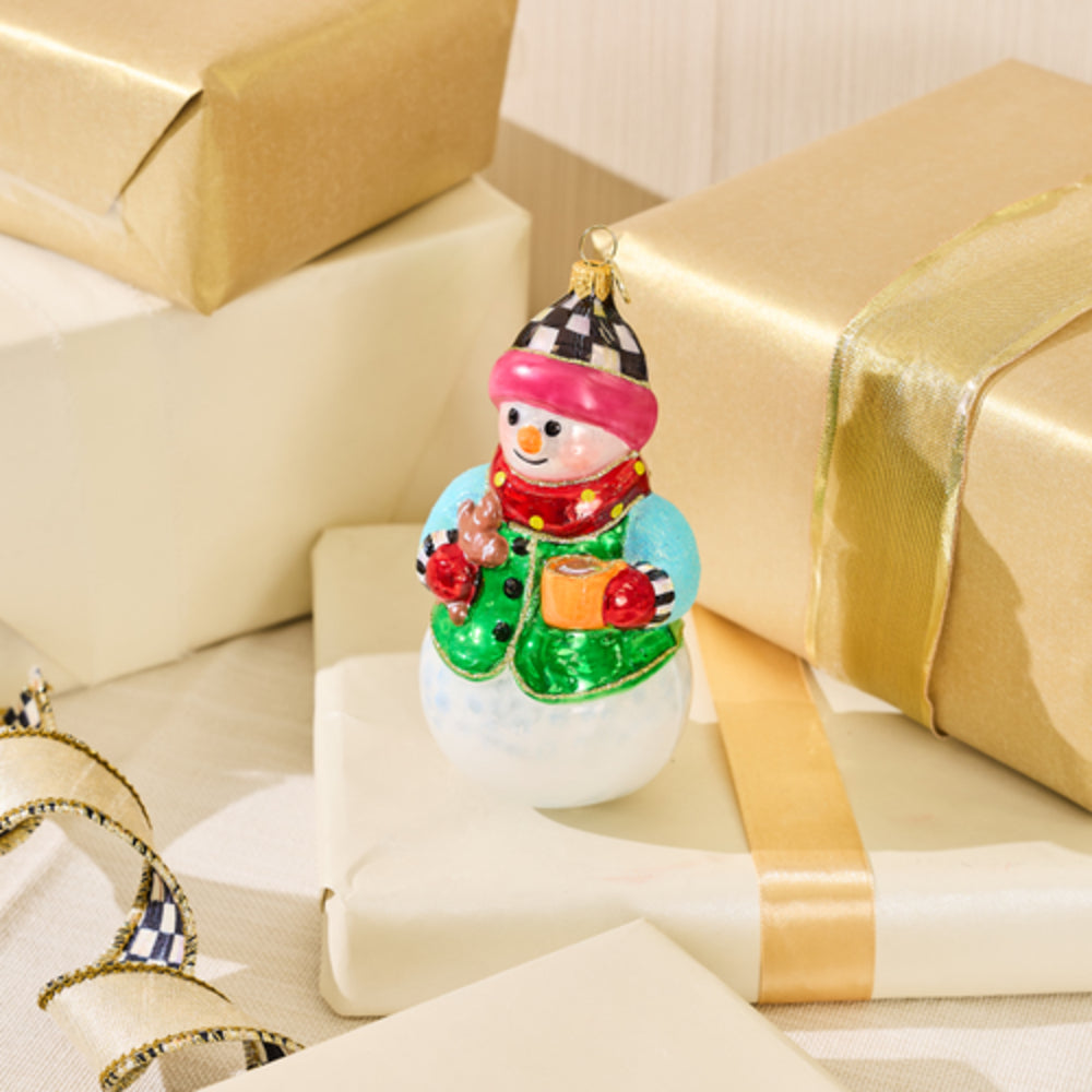 A small snowman Christmas ornament with a pink hat, red scarf, green coat, and blue sleeves, displayed on top of wrapped gift boxes with gold and cream paper and ribbons.