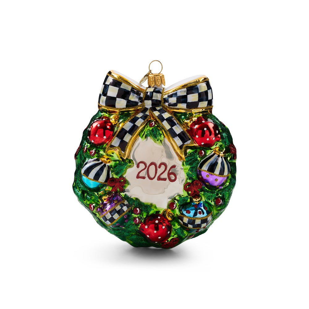 A Christmas ornament shaped like a green wreath with a red "2026" in the center, red and striped ornaments, and a black-and-white checkered bow at the top, shown on a plain white background.