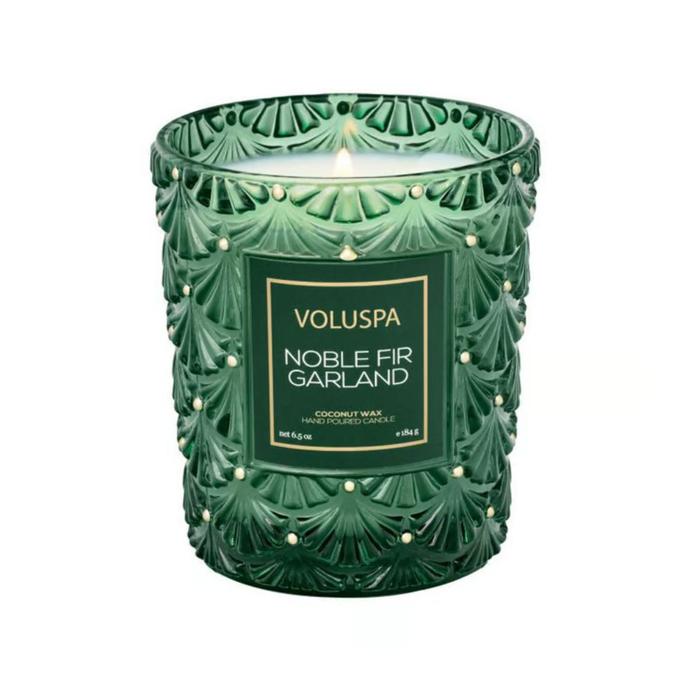 A green textured glass jar candle with a lit wick, featuring a front label that reads 'VOLUSPA NOBLE FIR GARLAND' on a plain white background.