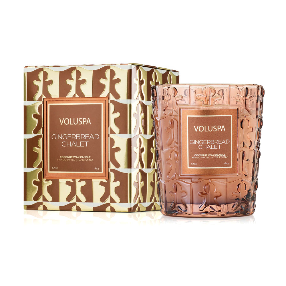 A VOLUSPA Gingerbread Chalet coconut wax candle in a textured rose-gold glass jar, shown next to its patterned brown and beige gingerbread-themed box on a white background.