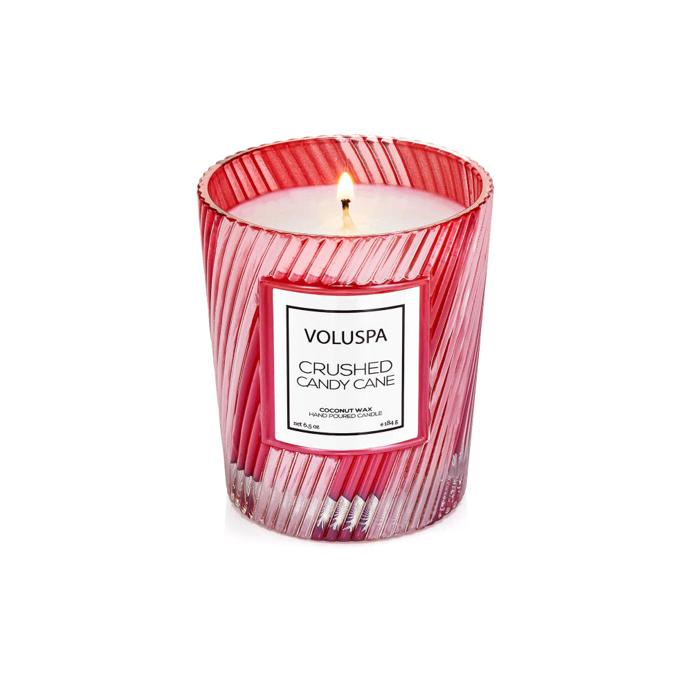 A lit Voluspa Crushed Candy Cane scented candle in a red and white striped glass jar, set against a plain white background.