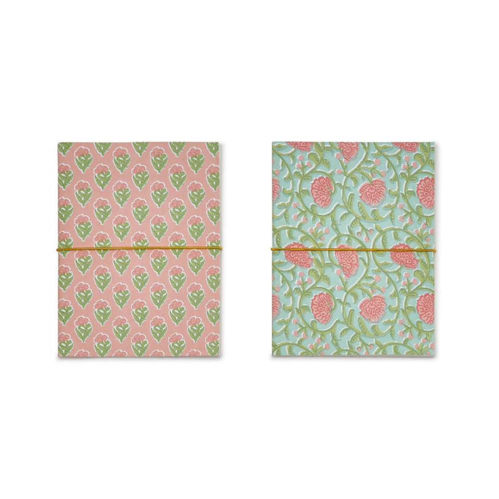 Two small patterned notebooks or journals side by side on a white background. The left one has a pink cover with a green floral pattern and a gold band across the middle.