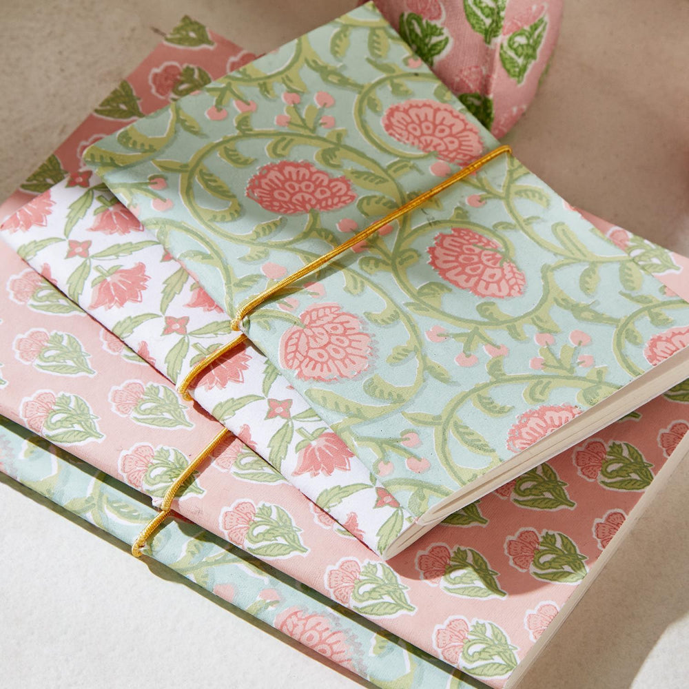 A stack of patterned notebooks or journals with a floral print in pink, sage green, and light blue, tied with thin yellow elastic bands, arranged on a neutral surface.