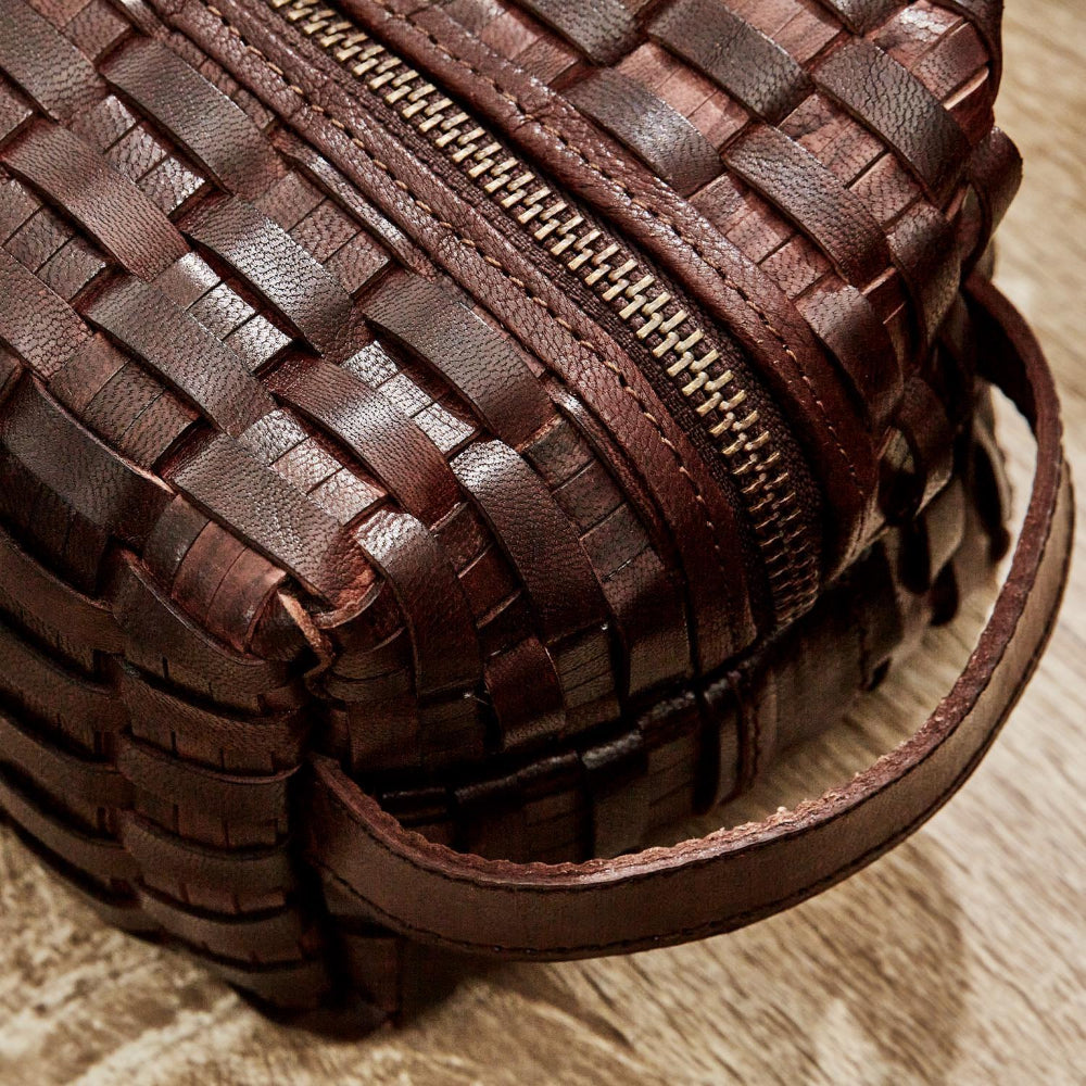 A close-up of a dark brown woven leather zippered pouch with a matching leather handle, resting on a light wood surface.