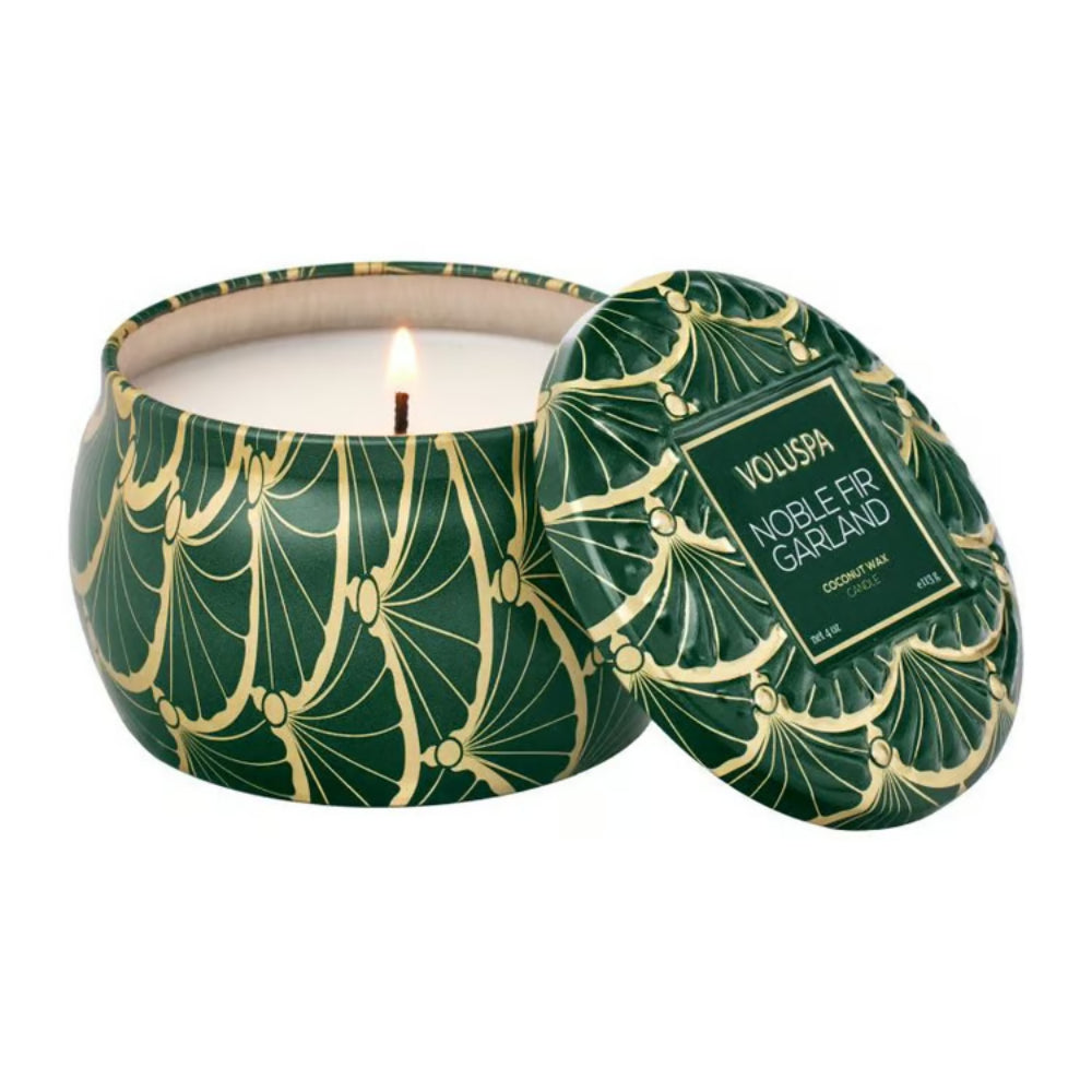 A green tin candle with gold decorative pattern with lid off and wick lit on a white background.