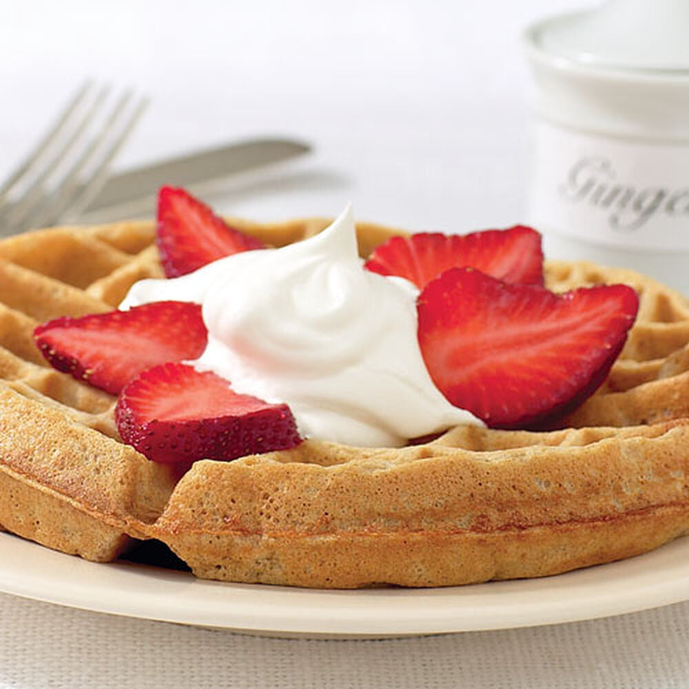 Waffle with strawberries and whipped cream on a plate, with a cup of coffee in the background.