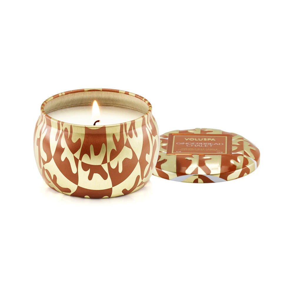 A lit candle in a round metal tin with a brown and beige camouflage pattern, shown next to its matching lid on a white background. The label on the lid reads 'VOLUMA COLD AIRY SCENTS'.
