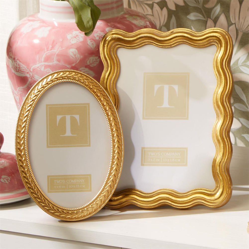A photo of a 5x7 gold wave edged picture frame on a white shelf with a pink vase and a gold edged oval picture frame and green floral wall paper in the backround.