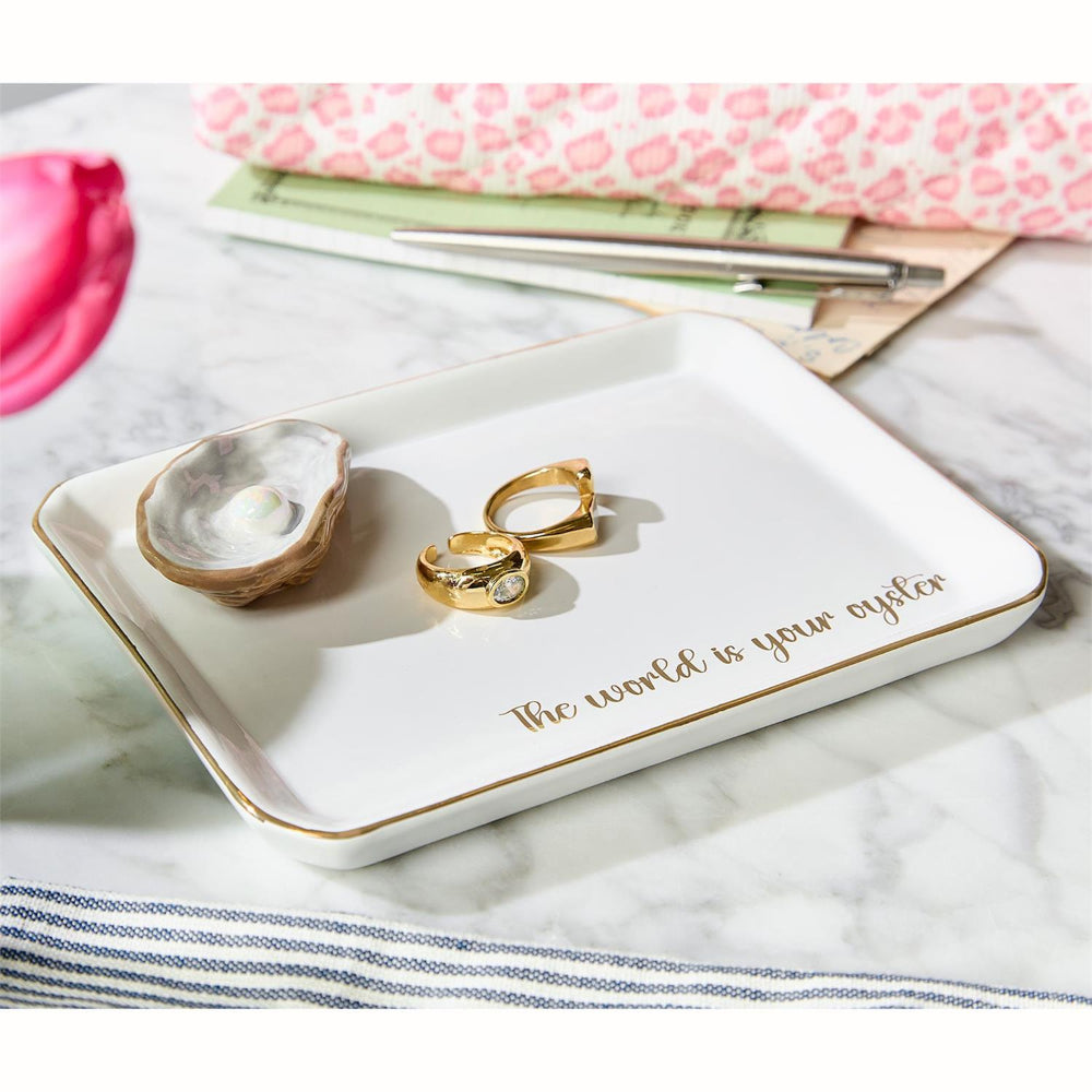 Jewelry tray with 'The world is your oyster' text, gold ring, and shell on a marble surface.