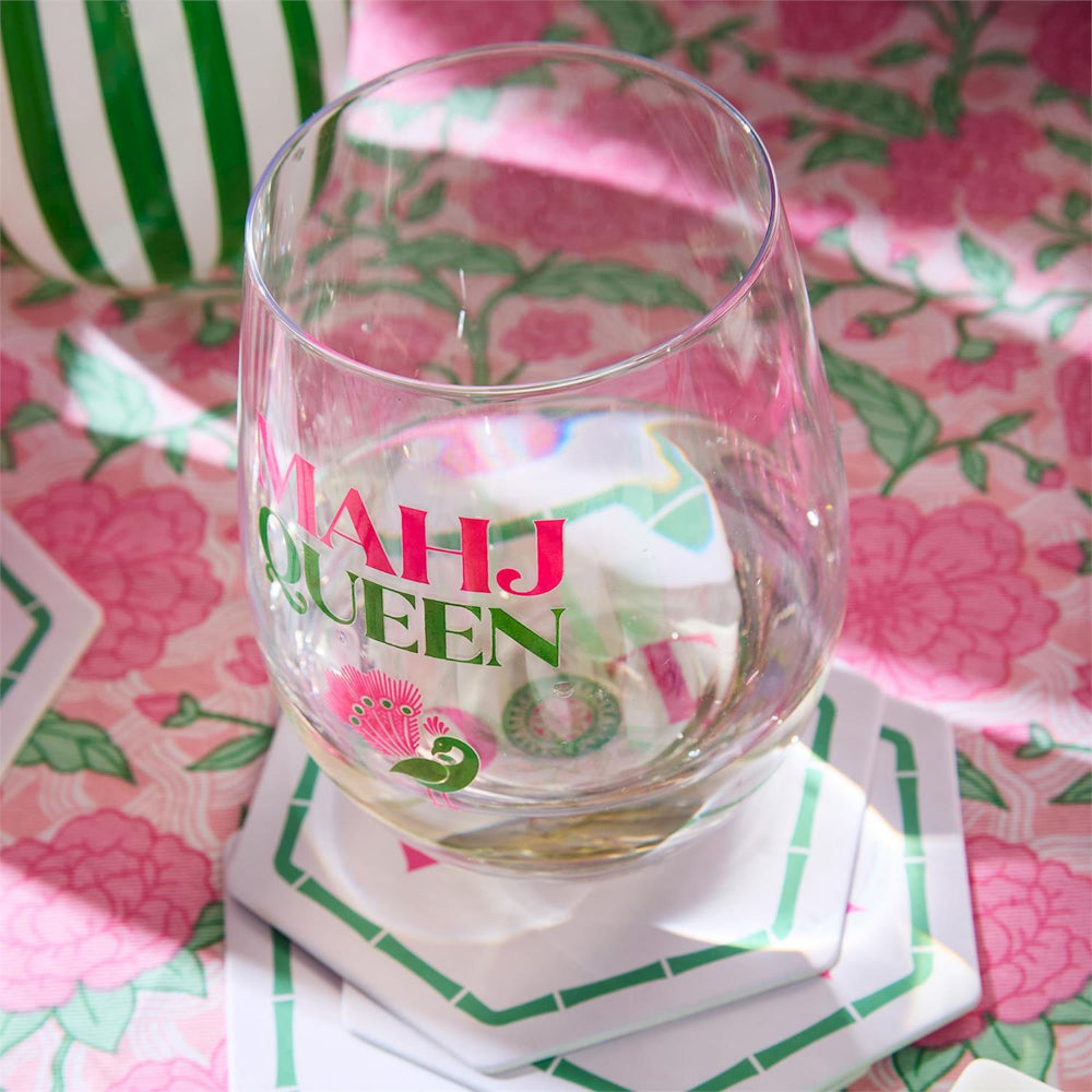 A clear stemless wine glass with pink and green printed text reading 'MAHJ QUEEN' sits on white coasters on a pink floral tablecloth.