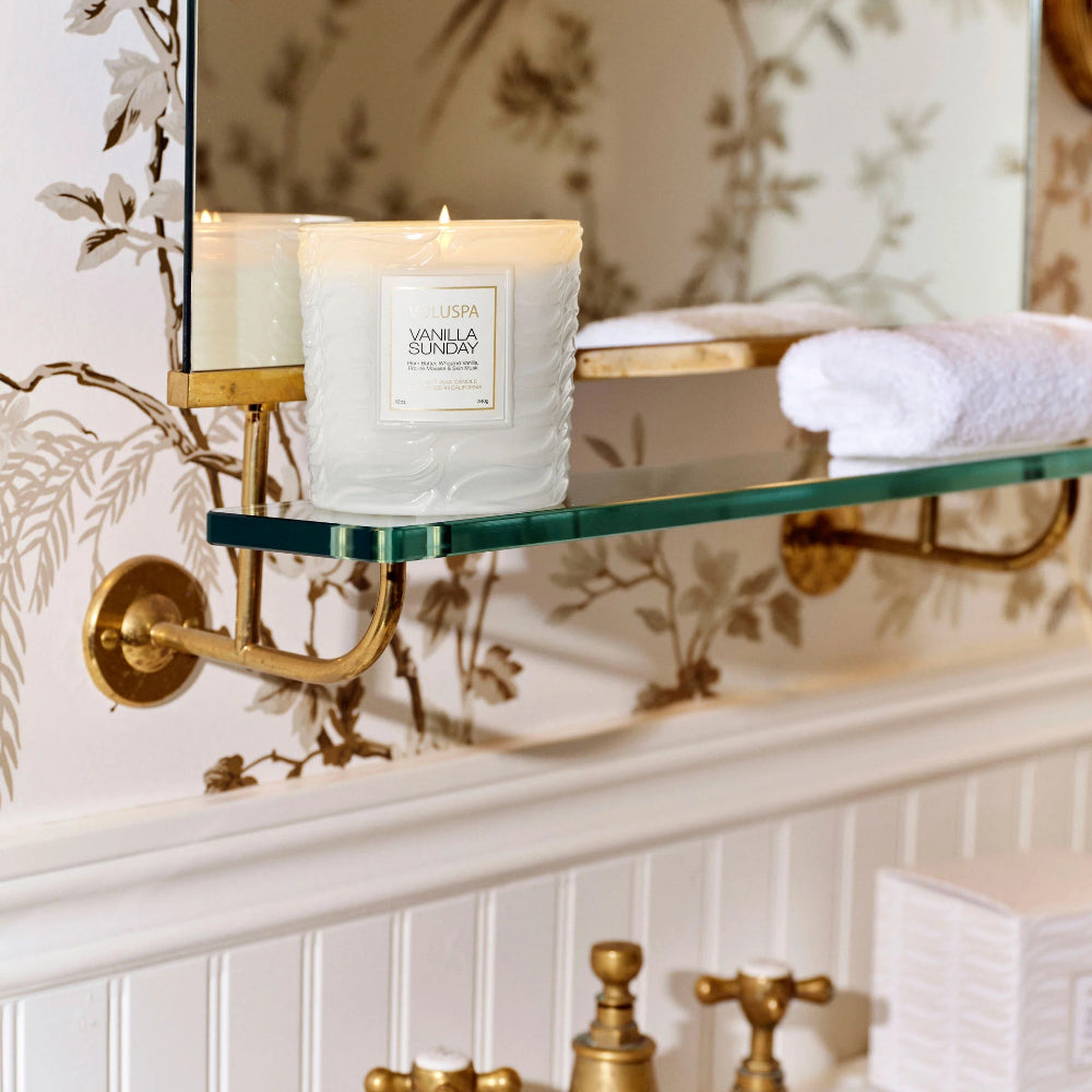 Candle on a glass shelf with floral wallpaper and gold accents.