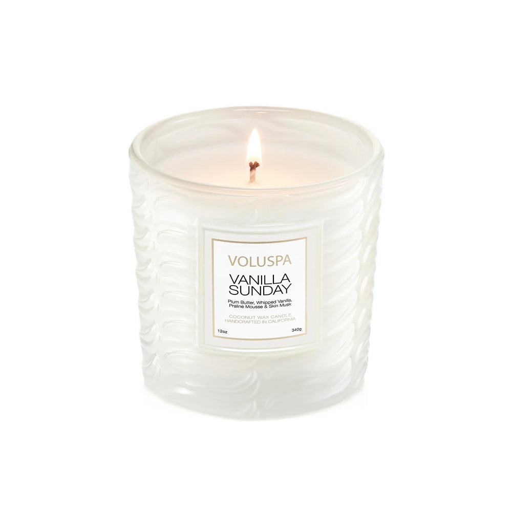 A lit Voluspa Vanilla Sunday scented candle in a textured, frosted white glass jar, shown against a solid white background.