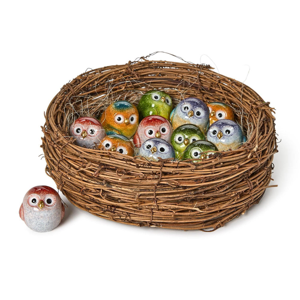A decorative woven twig nest filled with small painted owl figurines in assorted colors, with one owl figurine placed outside the nest on a white background.