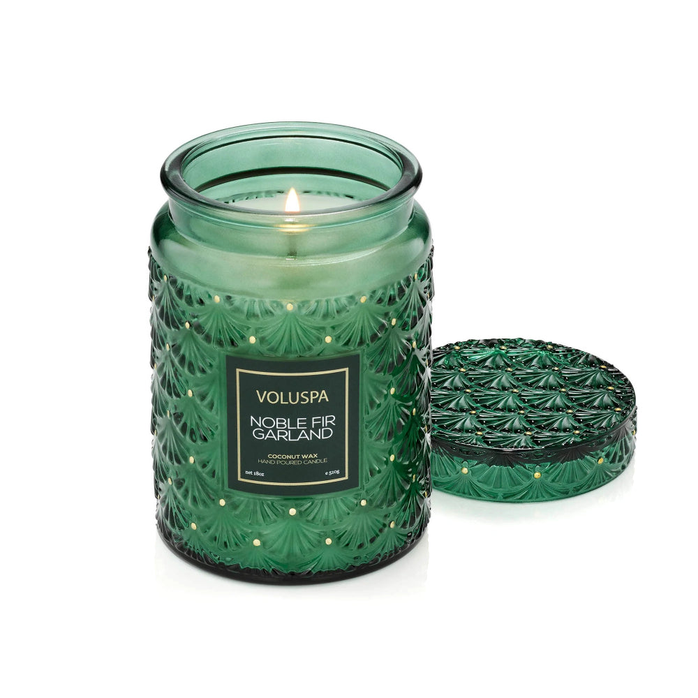 A green glass jar candle with a lit wick and matching textured lid, shown on a clean white background. The front label reads 'VOLUSPA NOBLE FIR GARLAND'.