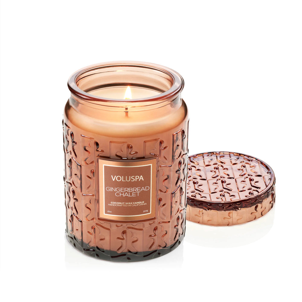 A lit Voluspa Gingerbread Chalet scented candle in a textured amber glass jar, with its matching lid resting beside it on a clean white background.
