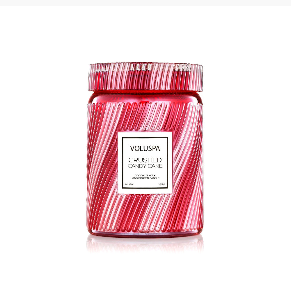 A Voluspa Coconut Wax candle in a clear glass jar with a red and white striped pattern resembling crushed candy canes, on a white background.