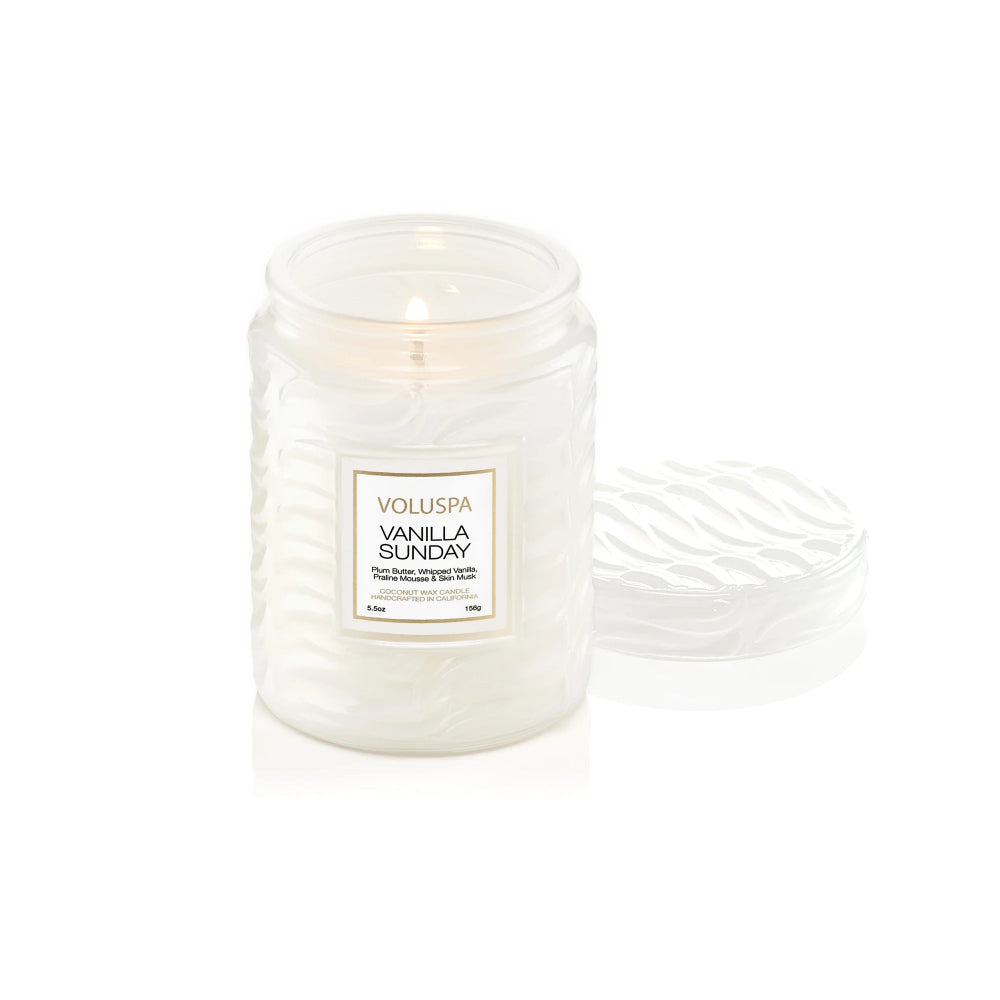 A Voluspa Vanilla Sunday scented candle in a textured clear glass jar with a white wax fill and a lit wick, shown next to its matching textured lid on a white background.