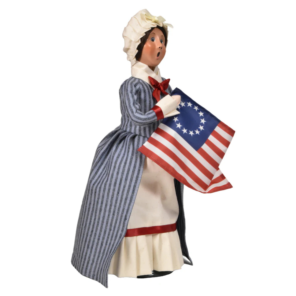 Right side photo of a figurine of a caroling Betsy Ross in historical attire holding an American flag on a white background