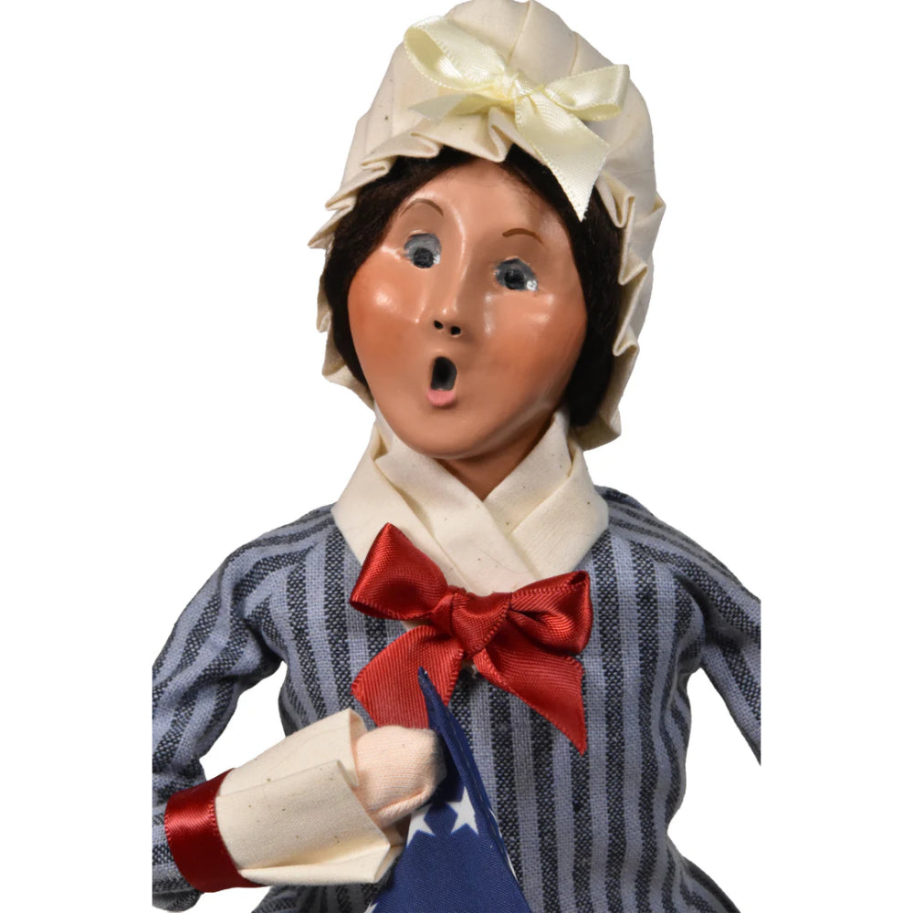 Close up of the face of a figurine of a caroling Betsy Ross in historical attire holding an American flag on a white background