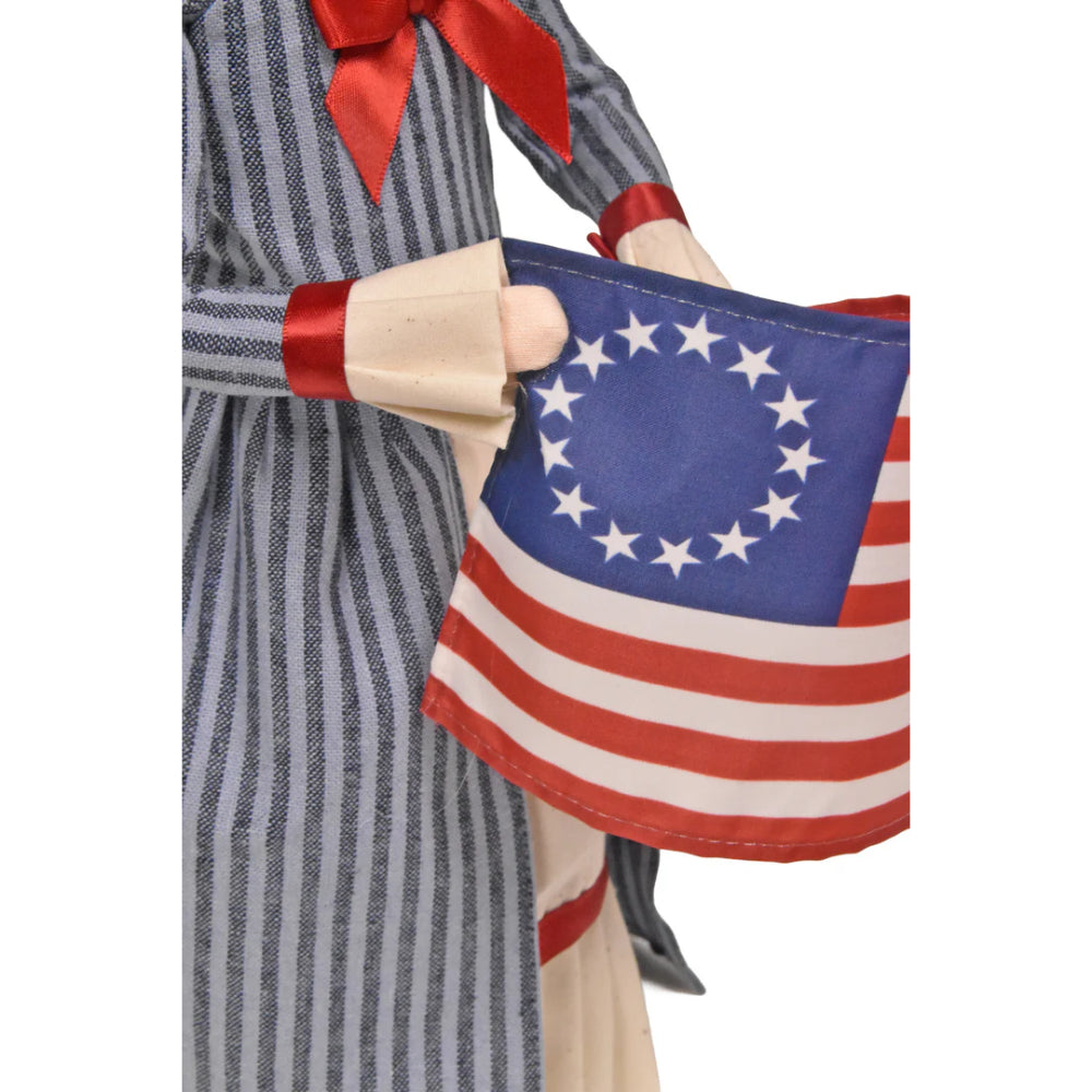An close up of the American flag held by a caroling figurine of Betsy Ross in historical attire holding an American flag on a white background