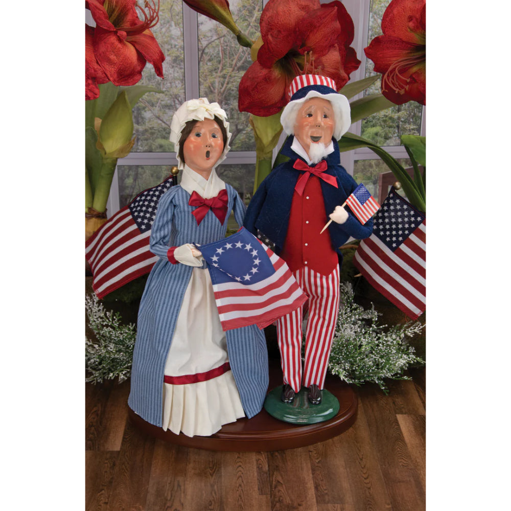 Decorative figurines of a caroling Betsy Ross and Uncle Sam holding American flags in a patriotic setting with red flowers and American flags