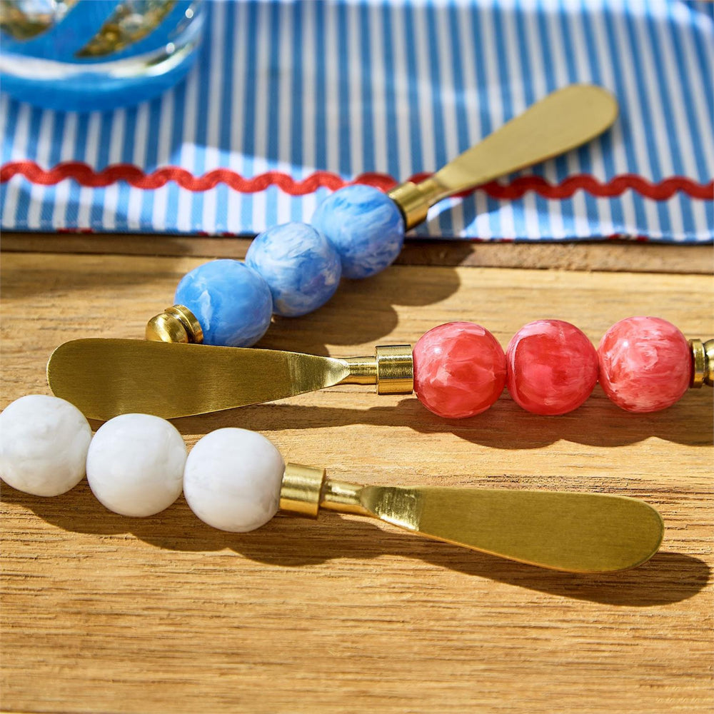 Three cheese knives with red, white and blue marble resin handles on a wooden surface, with a blue and white striped cloth in the background.