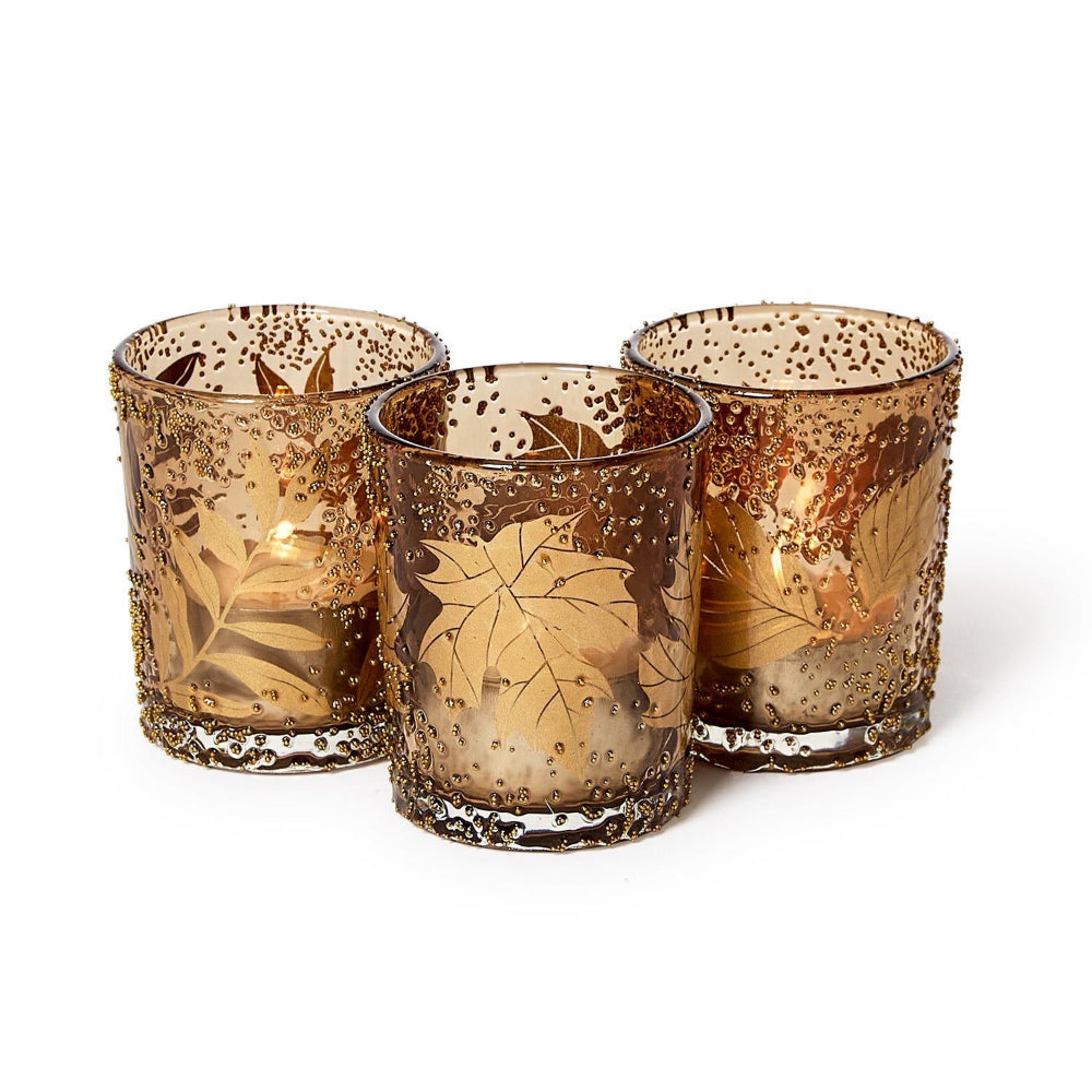 Three amber glass candle holders with gold leaf patterns and perforated speckled detailing, arranged on a white background.
