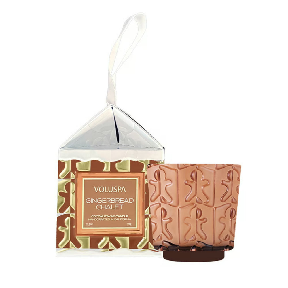 Voluspa Gingerbread Chalet scented wax candle ornament  in a clear gift box with a hanging loop, next to a small gingerbread-patterned candle holder on a white background.