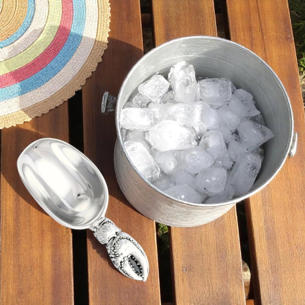 A silver metal ice bucket filled with ice cubes and a matching silver scoop with a lobster handle, placed on a wooden slatted surface outdoors.
