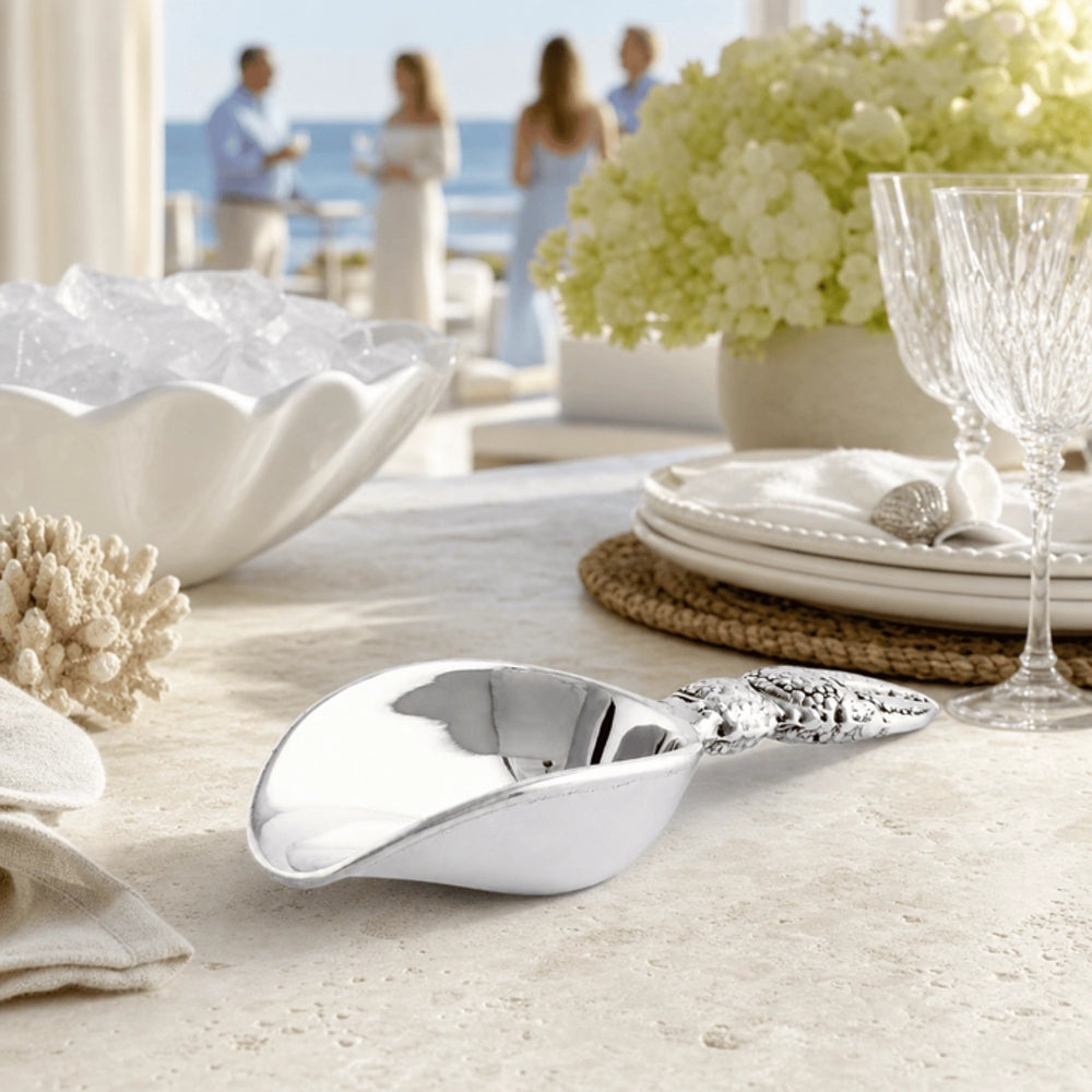 A polished silver serving scoop with an ornate handle resting on a light stone table, styled with white plates, a glass, and a bowl of ice in a bright, upscale outdoor setting with people blurred in the background.