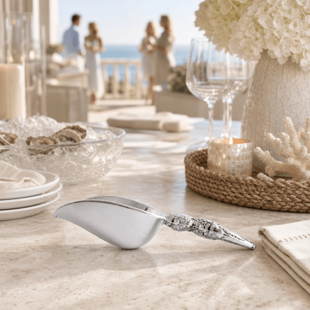 A polished silver ice scoop with an ornate handle resting on a light stone table in a beautifully styled outdoor dining setting with blurred figures in the background.