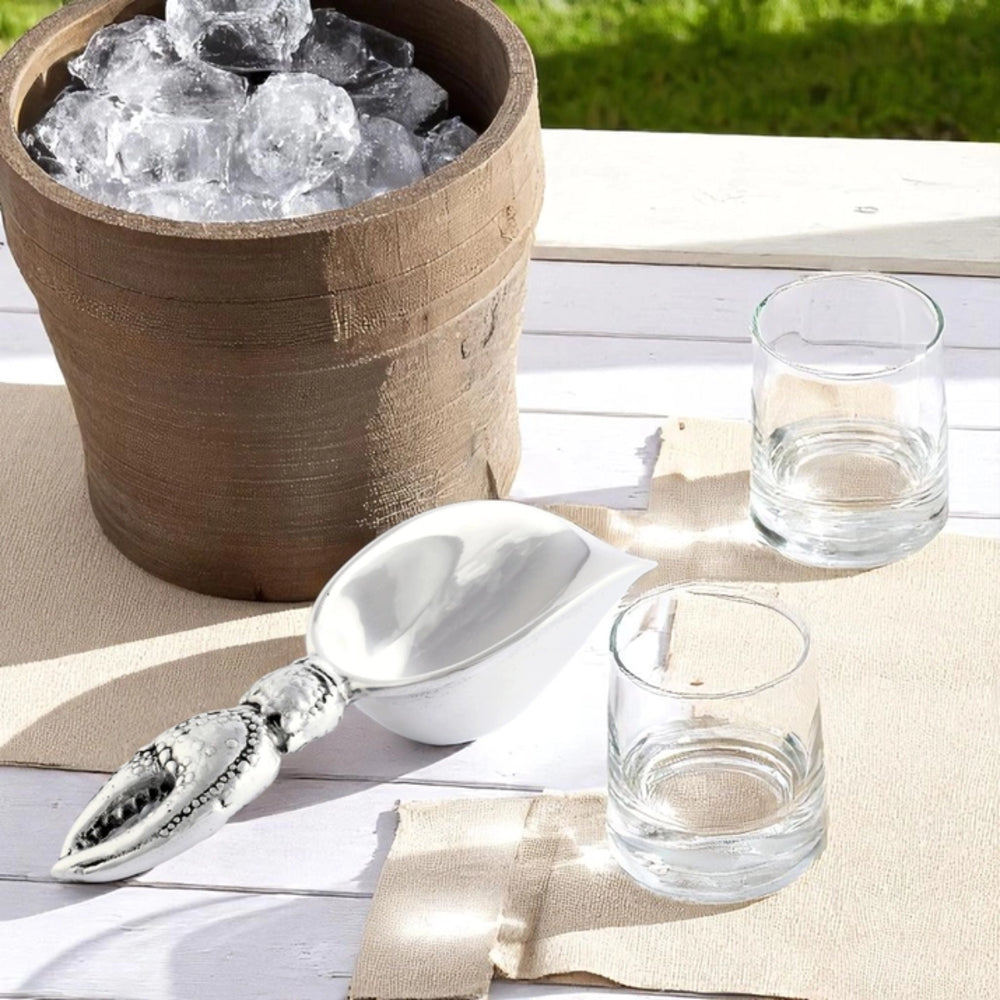 A silver metal ice scoop resting on a white wooden table beside a rustic wooden bucket filled with ice, with two clear drinking glasses nearby on a beige cloth.