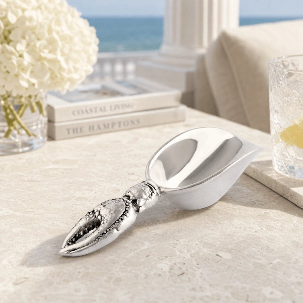 A silver lobster claw scoop with a polished bowl, resting on a light stone table in a coastal-themed setting with a vase of white flowers, books, and a glass of lemonade in the background.