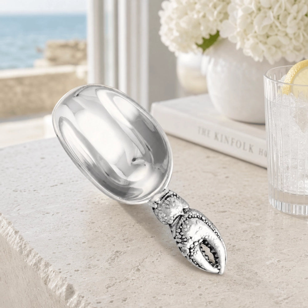 A polished silver lobster scoop with a detailed lobster claw handle, resting on a light stone countertop in a bright kitchen or bar setting.