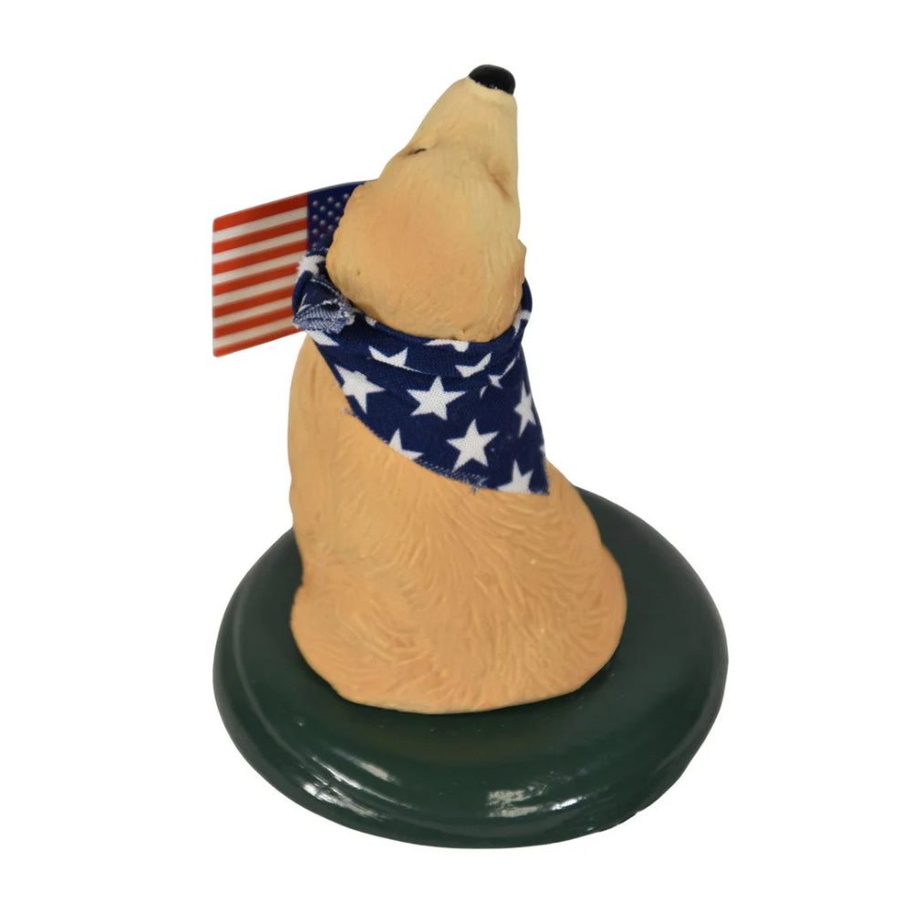 Back of patriotic golden retriever  figurine wearing a American flag scarf  and holding an American flag on a green base on a white background