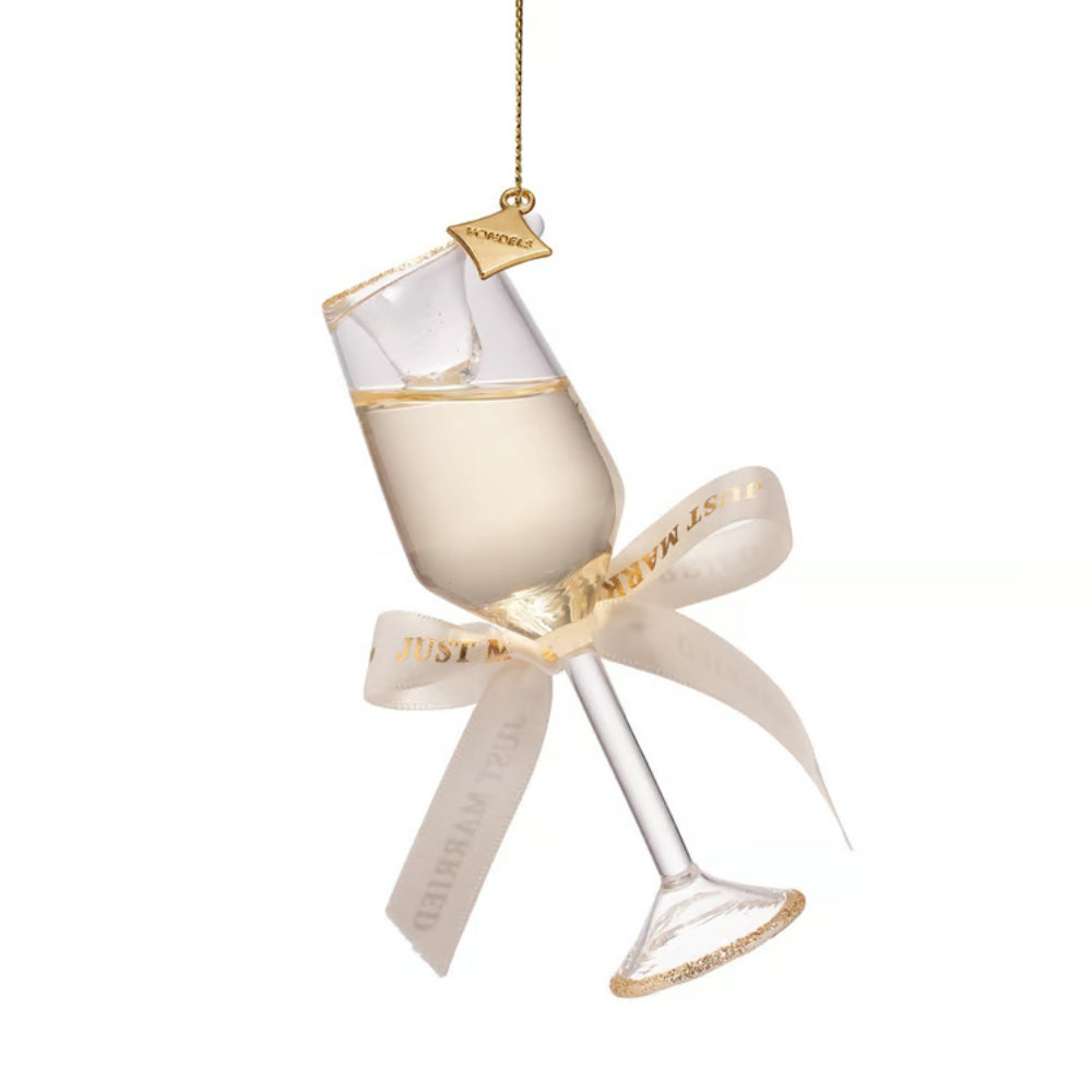 A clear glass champagne flute ornament with gold trim, filled with pale champagne-colored liquid, tied with a cream ribbon printed with gold lettering, hanging from a gold string on a white background.