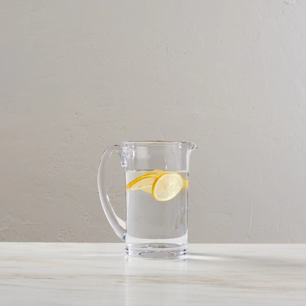 A clear glass pitcher filled with water and lemon slices, set on a light marble surface against a neutral textured wall.