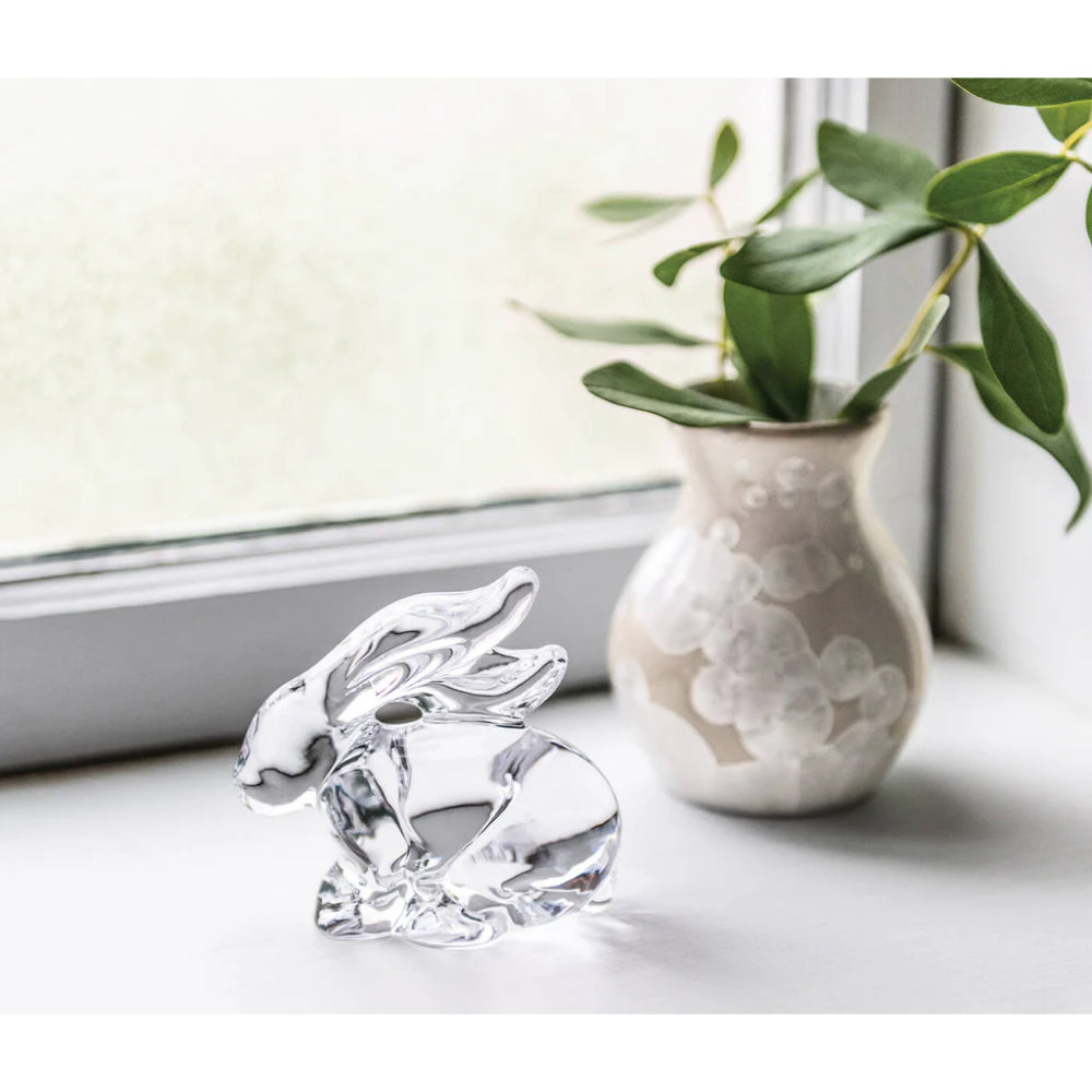Crystal rabbit figurine next to a ceramic vase with greenery on a light surface.