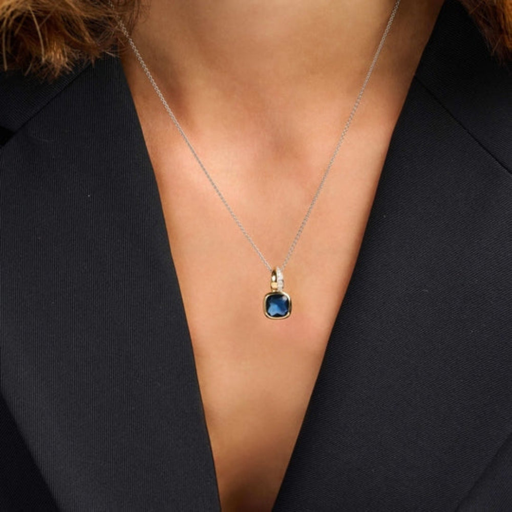 A close-up of a model wearing a silver chain necklace with a gold bezel-set oval blue gemstone pendant, styled with a black blazer.