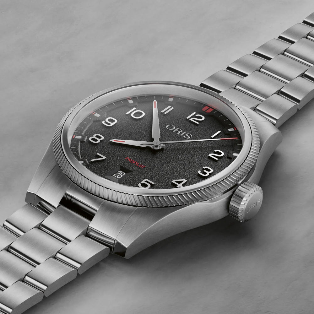 An Oris stainless steel watch with a black textured dial, silver-tone case, and matching metal link bracelet, photographed on a neutral gray surface.