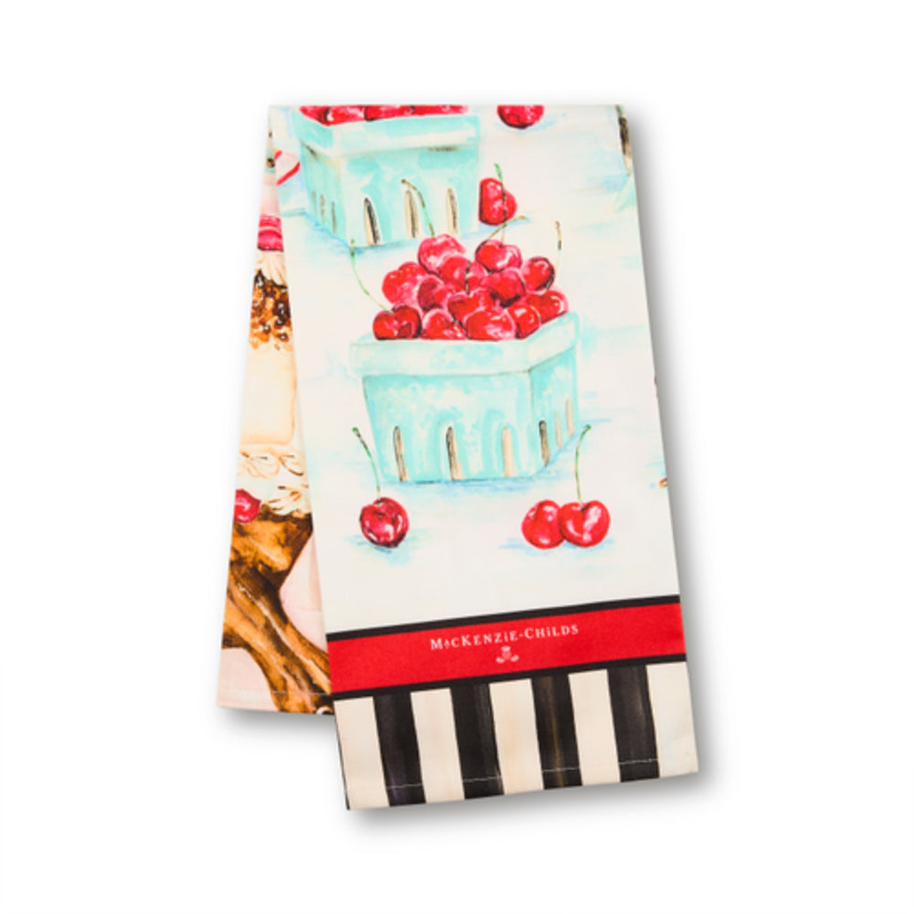 A kitchen towel featuring a vintage-style illustration of light blue baskets filled with red cherries, with a red band reading 'MICKENIE-CHILDS' and black-and-white piano keys at the bottom, shown on a white background.